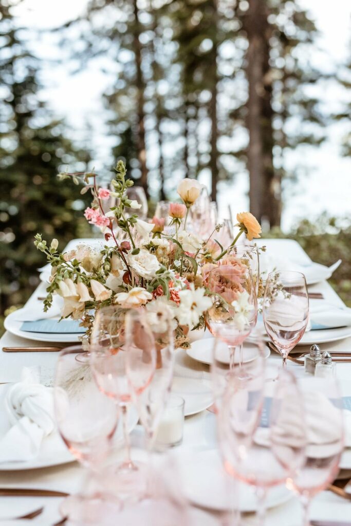 Elegant outdoor table setting with pink-tinted glassware, white plates, and a floral centerpiece featuring various pastel flowers. The background shows blurred trees and hints of water.