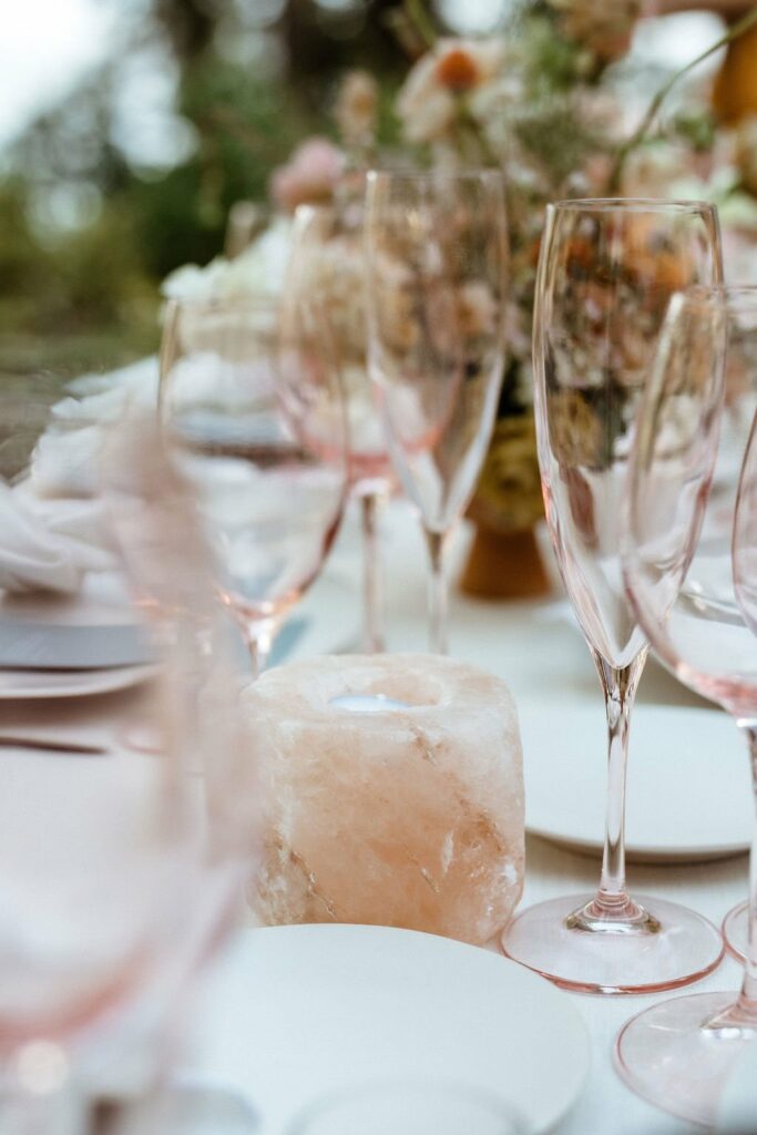 A close-up of a table setting featuring empty glasses, white plates, and a pinkish salt candle holder. A blurred floral arrangement is in the background.