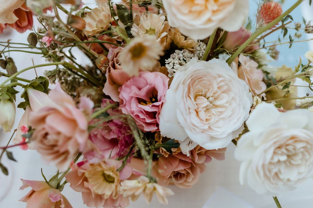 A close-up of a bouquet featuring various flowers, including white, pink, and peach roses, and other assorted blooms and greenery, all arranged in a natural, organic style.