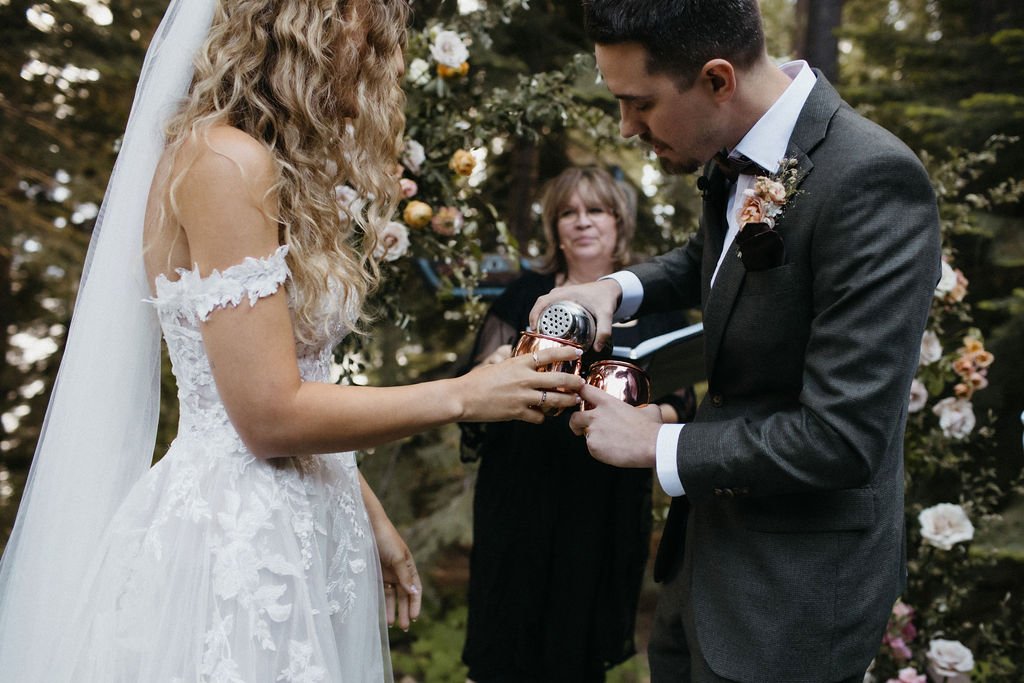 A bride and groom pour a drink together into copper mugs during an outdoor wedding ceremony, with an officiant and floral decorations in the background.