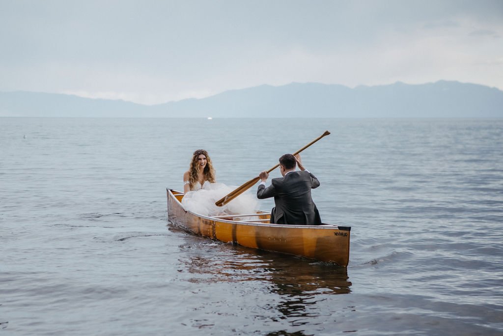 A couple dressed in wedding attire paddles a wooden canoe on a calm lake with mountains in the background.