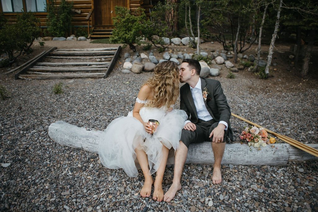 A couple sits on a log, barefoot, with the woman in a wedding dress and the man in a suit, sharing a kiss. They are outdoors with a cabin and greenery in the background.