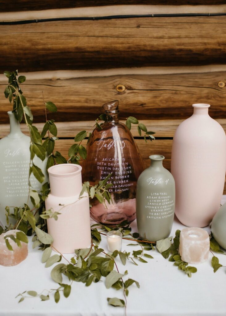 Various pastel-colored vases with handwritten names and seating assignments displayed on a white cloth, accented with greenery and small candles, set against a rustic wooden backdrop.