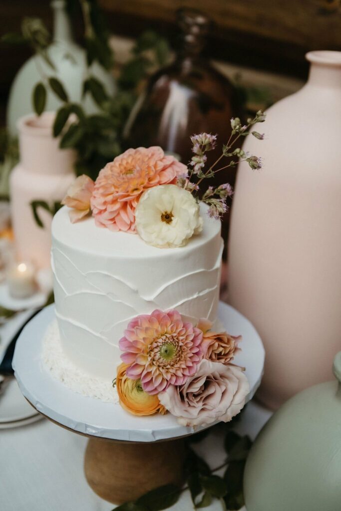 A white tiered cake decorated with pink and peach flowers is displayed on a wooden stand, with various pastel-colored vases and greenery in the background.