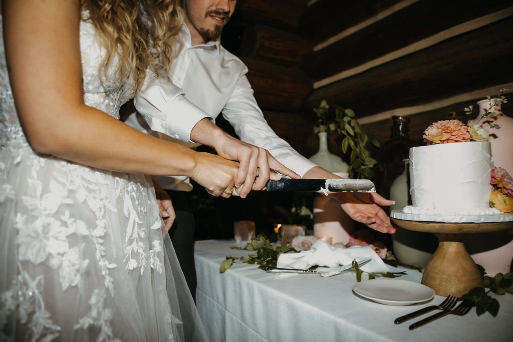 A couple cuts a white tiered wedding cake together with a knife. The table is decorated with flowers, greenery, napkins, and plates.