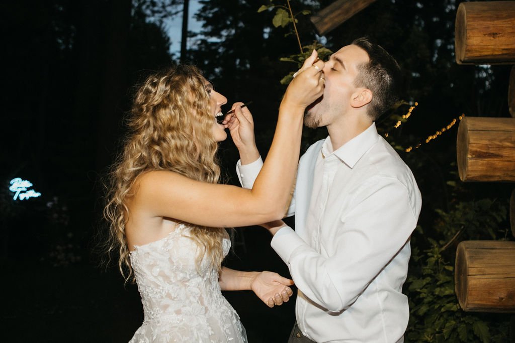 A bride and groom are feeding each other cake outdoors, with the bride wearing a white dress and the groom in a white shirt.