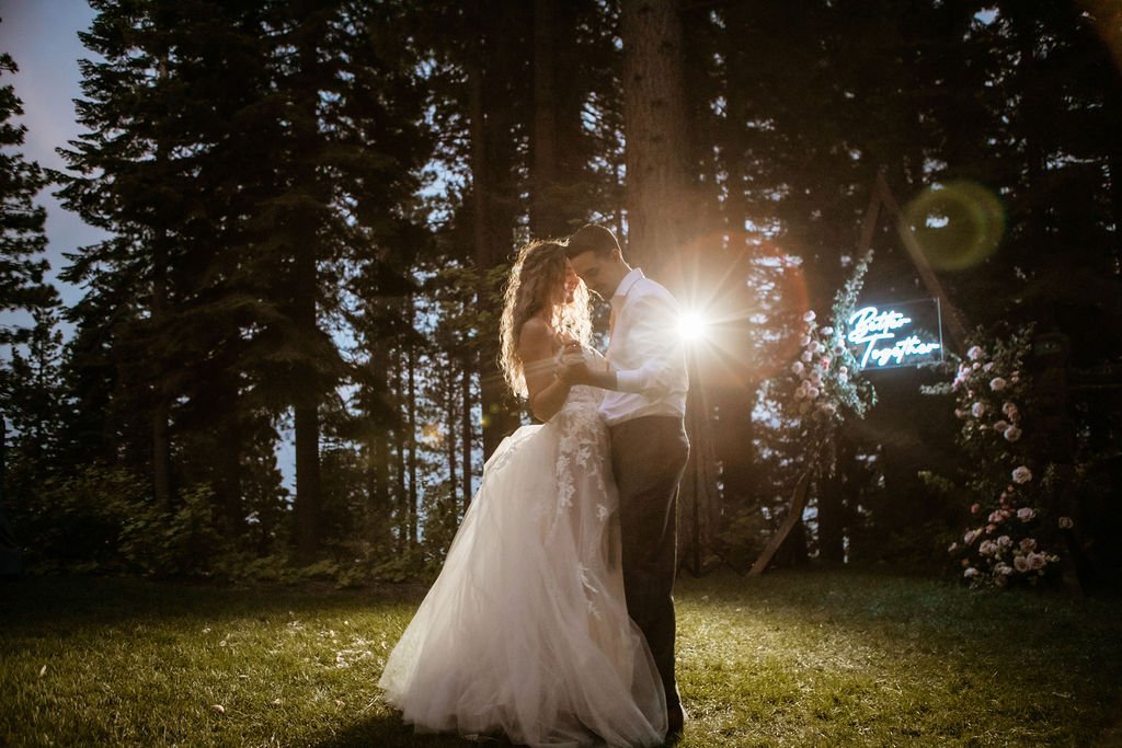 A bride and groom share a dance at an outdoor evening wedding surrounded by trees, with soft lighting and a decorative floral arrangement in the background.