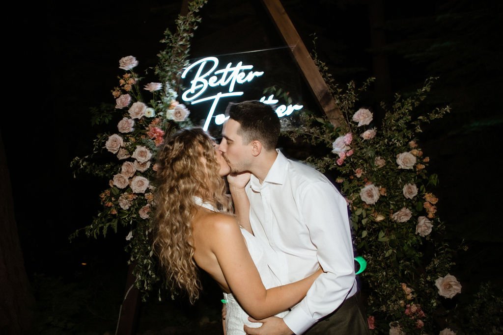 A couple shares a kiss in front of a floral arrangement and a neon sign that reads "Better Together".