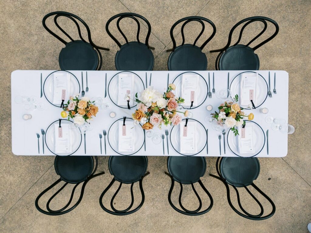 A rectangular table with white tablecloth, set for eight with black chairs, plates, menus, and floral centerpieces of white and peach flowers. The table setup is viewed from above.