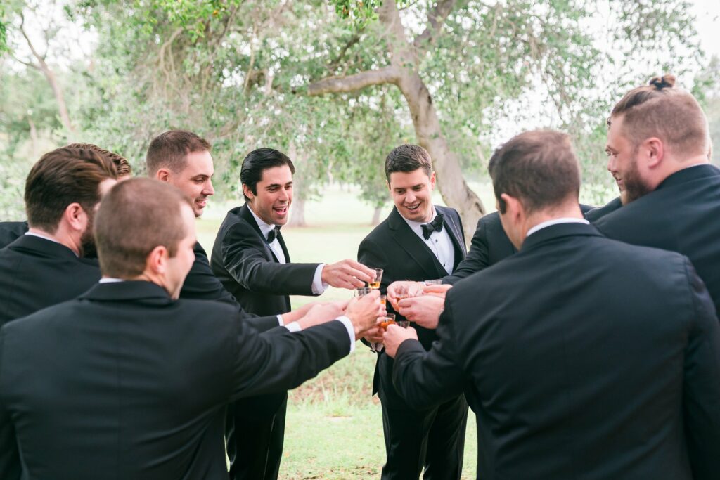 A group of men in black suits and bow ties stand in a circle outside, raising glasses in a toast. Trees and grass are visible in the background.