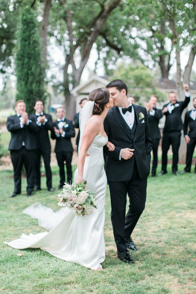 A bride and groom share a kiss outdoors, surrounded by a cheering wedding party dressed in black suits and ties. The bride holds a bouquet and wears a white gown, while the groom is in a black tuxedo.