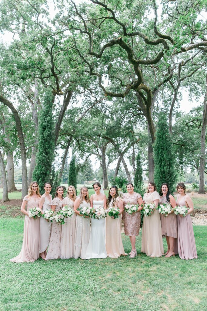 A group of twelve women standing outdoors in a wooded area, all dressed in light, pastel-toned dresses, holding bouquets of white and green flowers.