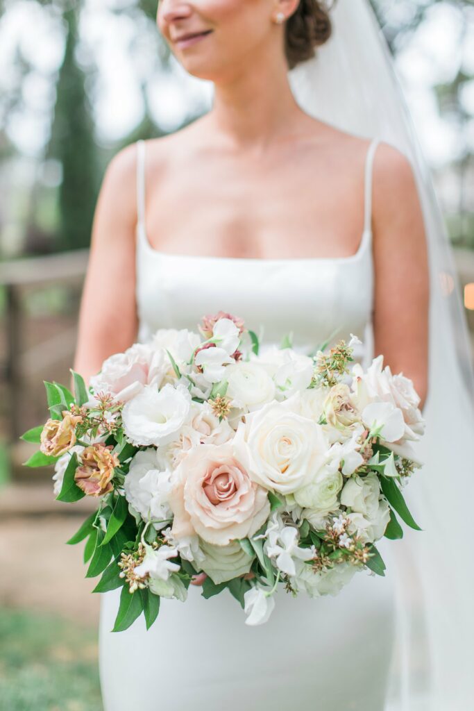A bride in a white dress holds a bouquet of white and blush pink flowers with greenery.