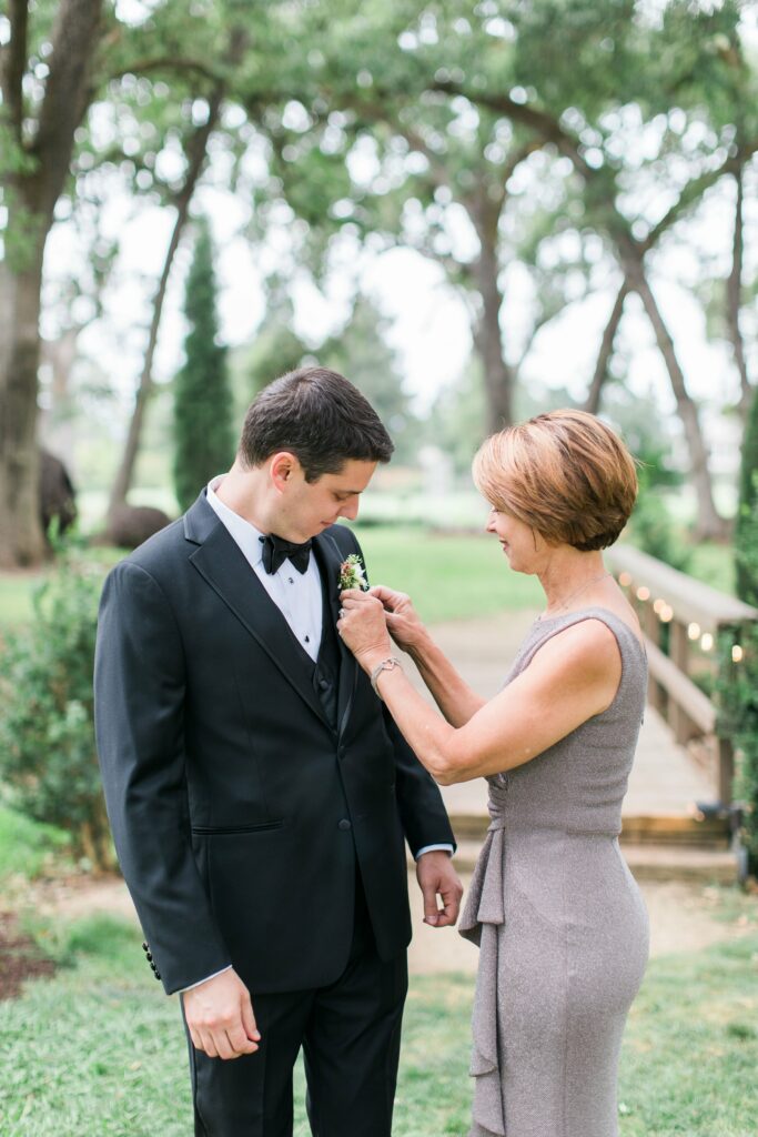 A woman in a gray dress adjusts the boutonniere on a man in a black tuxedo, outdoors in a park-like setting.