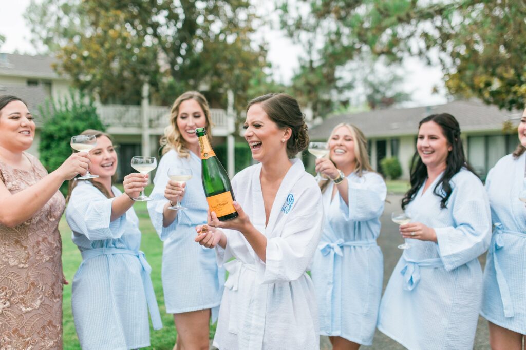 A group of women in light blue robes and one in a brown dress are outside celebrating with champagne, smiling and raising glasses. One woman is holding a champagne bottle. Trees and buildings are in the background.