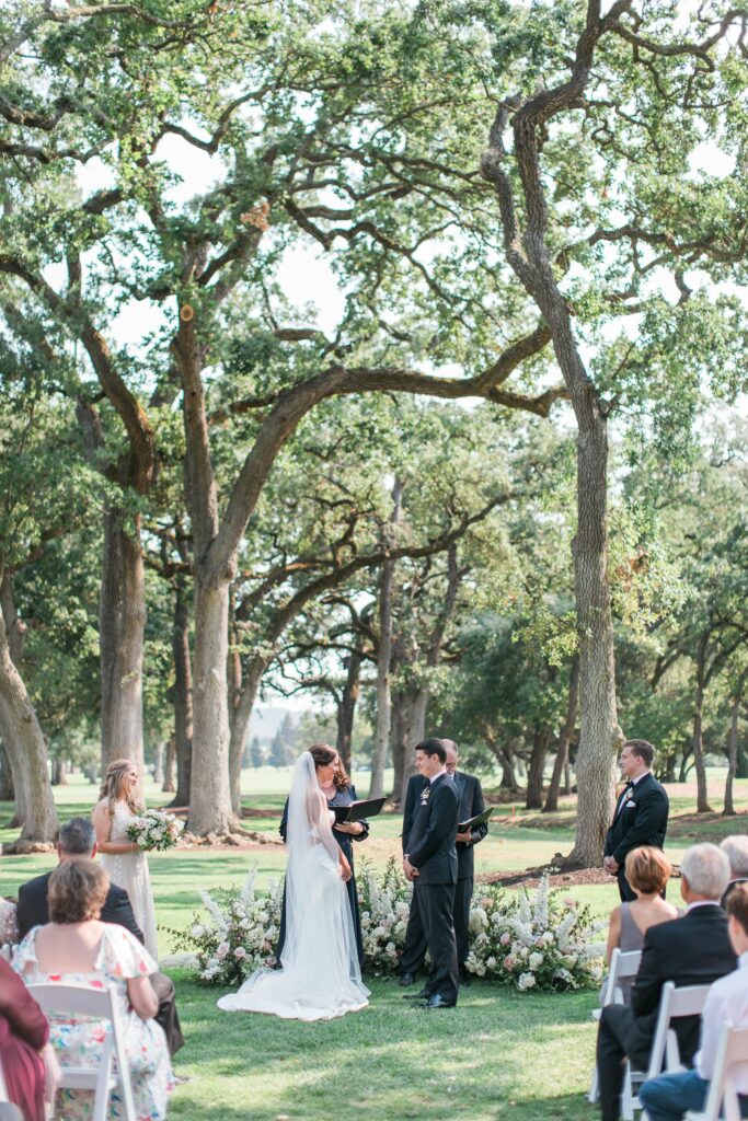 A couple stands facing each other, holding hands during an outdoor wedding ceremony beneath tall trees, surrounded by guests seated in rows, with white floral arrangements in the background.
