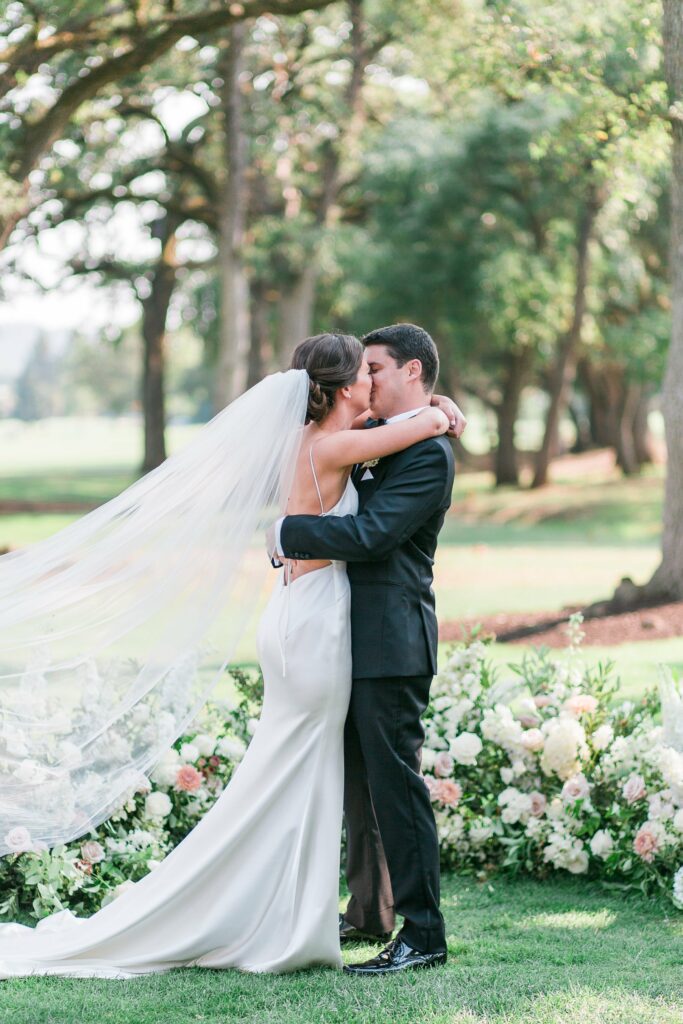 A bride and groom share a kiss outdoors, surrounded by flower arrangements, with trees and greenery in the background. The bride wears a white dress with a long veil, and the groom wears a black suit.