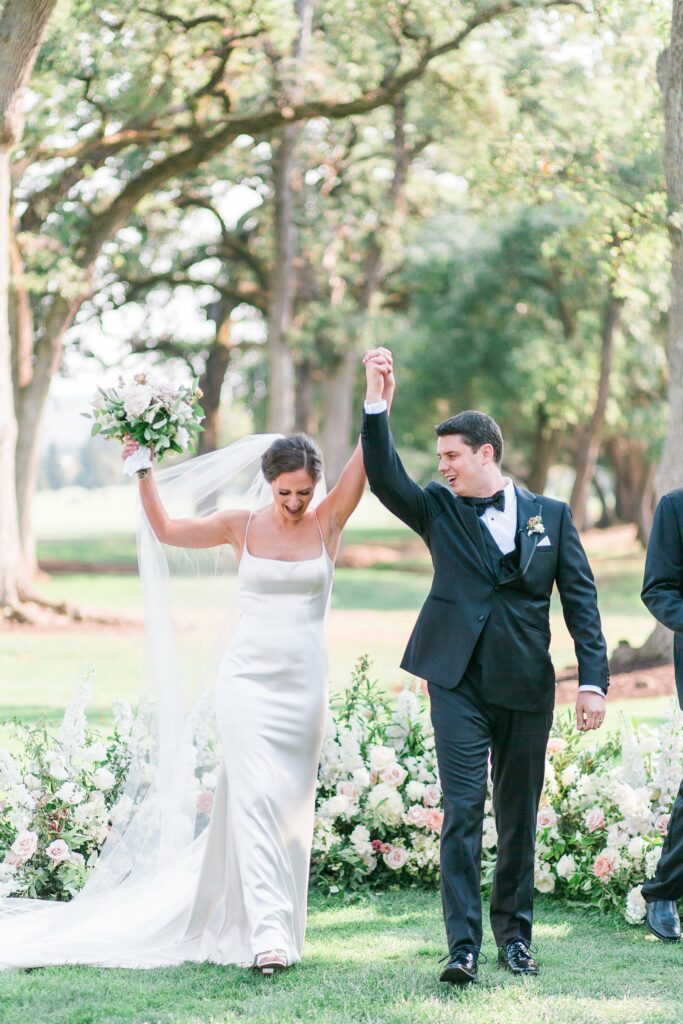 A bride and groom celebrate outdoors, holding hands raised in the air. The bride holds a bouquet and wears a long white dress. The groom wears a black tuxedo. Trees and flowers are in the background.