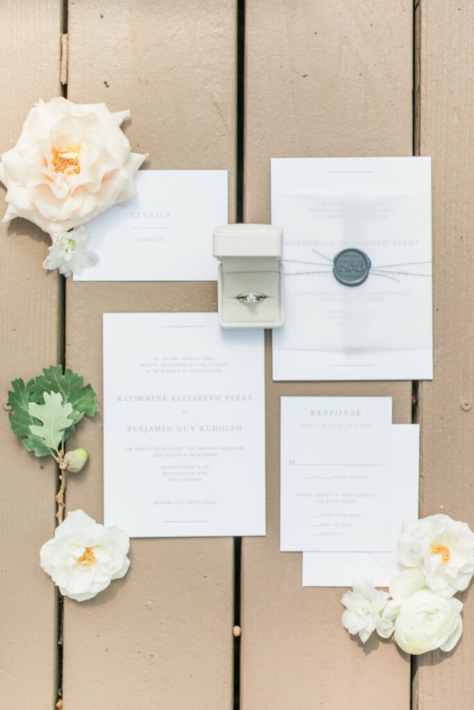 Wedding invitations and response cards are arranged on a wooden surface with white flowers. An open ring box displaying an engagement ring and a wax-sealed envelope are also visible.