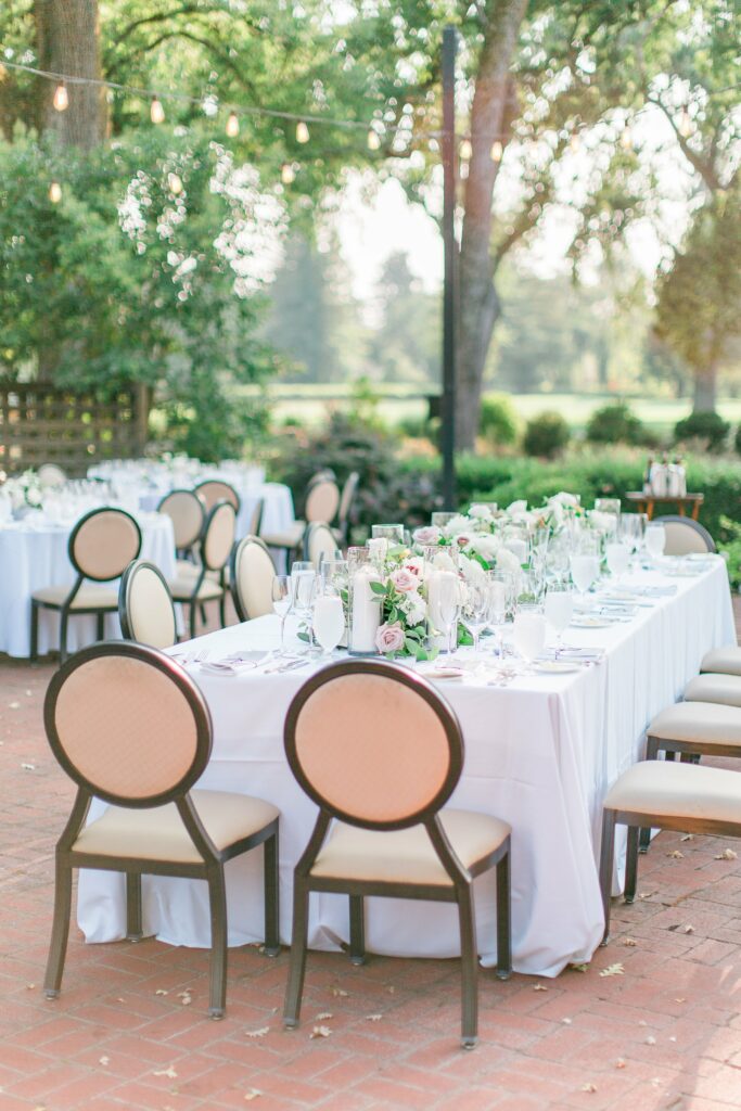 Outdoor dining setup with white tablecloths, floral centerpieces, glassware, and round-back chairs in a garden setting. String lights overhead.