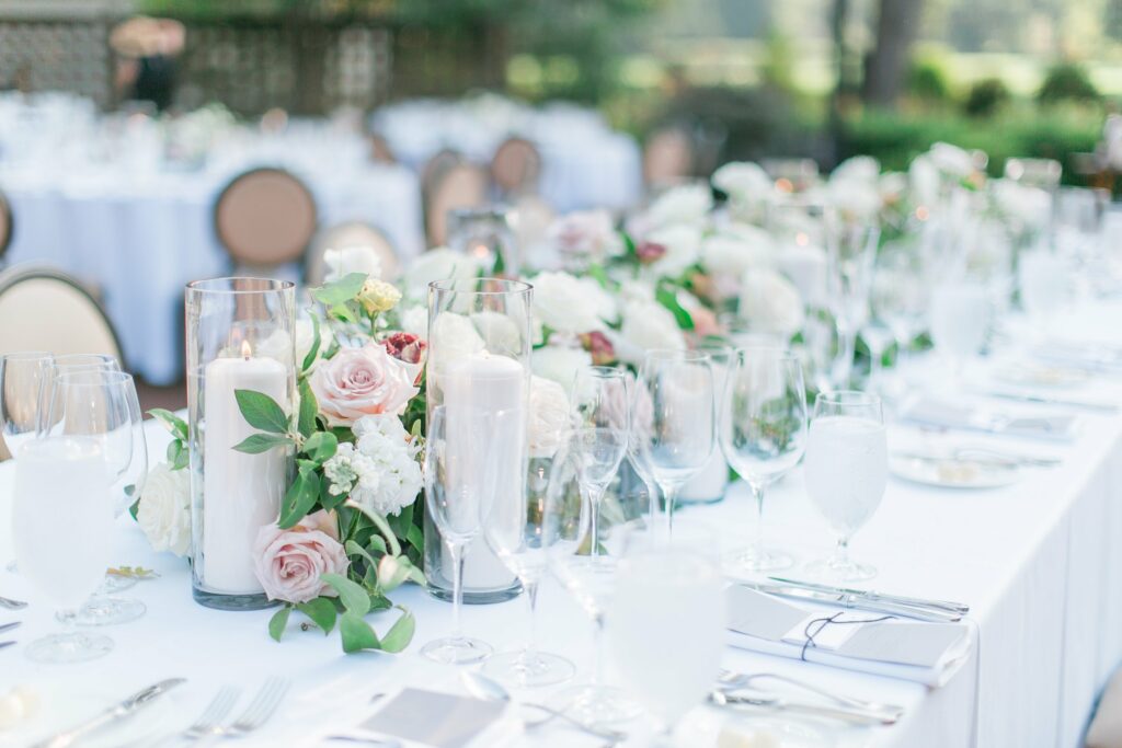 A long table set for a formal event features white tablecloths, floral centerpieces with pink and white flowers, candles, and neatly arranged glassware and cutlery.