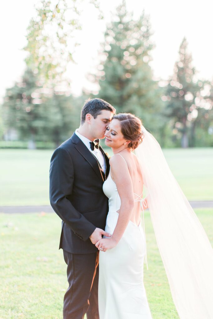 A bride in a white dress and veil stands holding hands with a groom in a black suit and bow tie outdoors on a sunny day, with trees in the background.