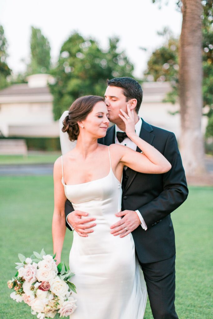 A bride in a white dress holding a bouquet of flowers is embraced by a groom in a black suit and tie who kisses her on the forehead in an outdoor setting.