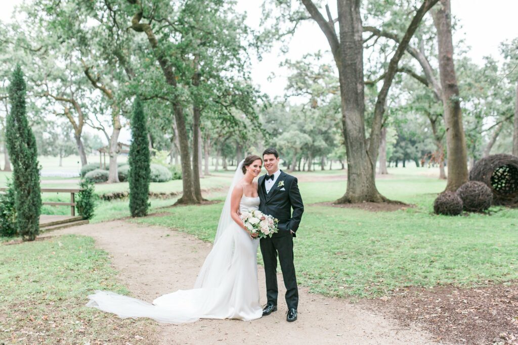 A bride in a white dress and a groom in a black tuxedo stand together on a path in a lush green park with trees in the background. The bride holds a bouquet of flowers.