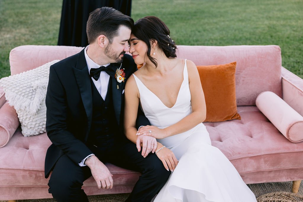 A couple dressed in wedding attire sit closely on a pink sofa outdoors. The bride wears a white gown, and the groom is in a black suit. They lean in with their foreheads touching, holding hands.