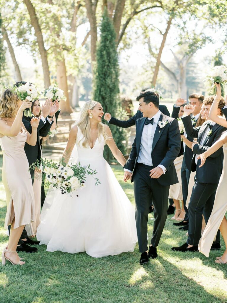 A bride and groom, holding hands and smiling, walk through a group of cheering wedding party members outdoors. The bride is in a white gown, and the groom is in a dark suit.