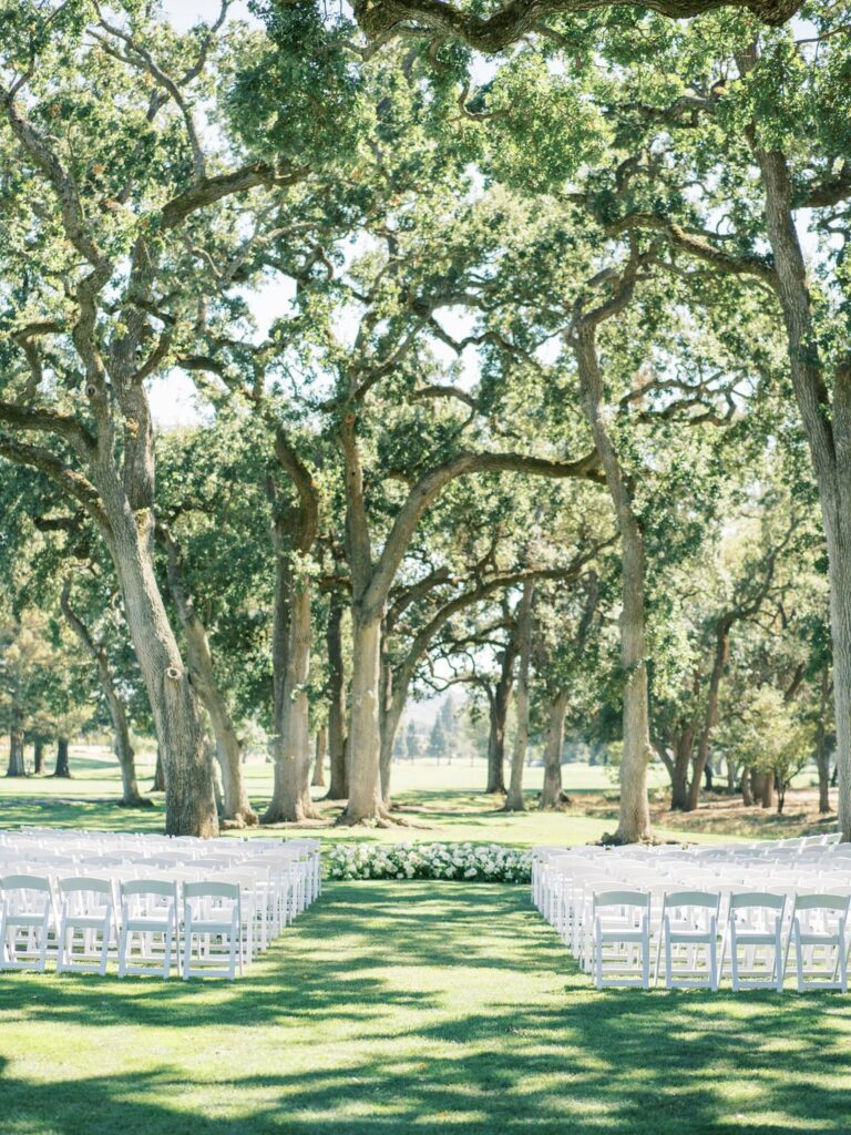 Rows of white chairs are set up outdoors in a wooded area with tall trees, forming an aisle toward a lush green forest backdrop.
