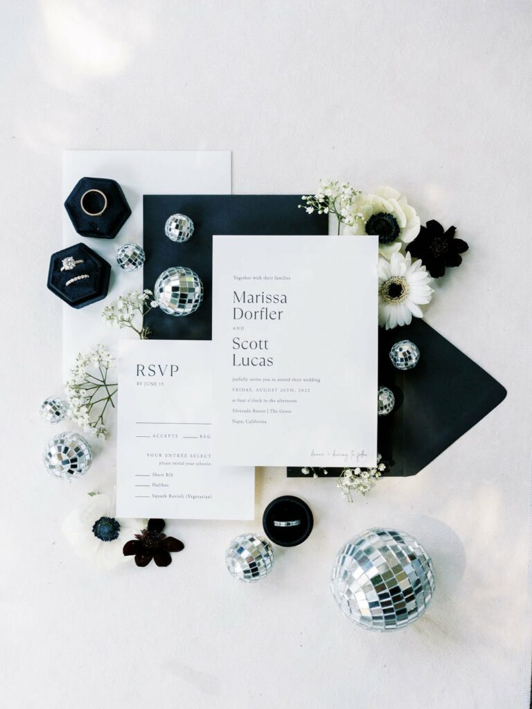 Elegant wedding invitation suite with black envelopes, RSVP card, rings in black boxes, white flowers, and small disco balls as decor.