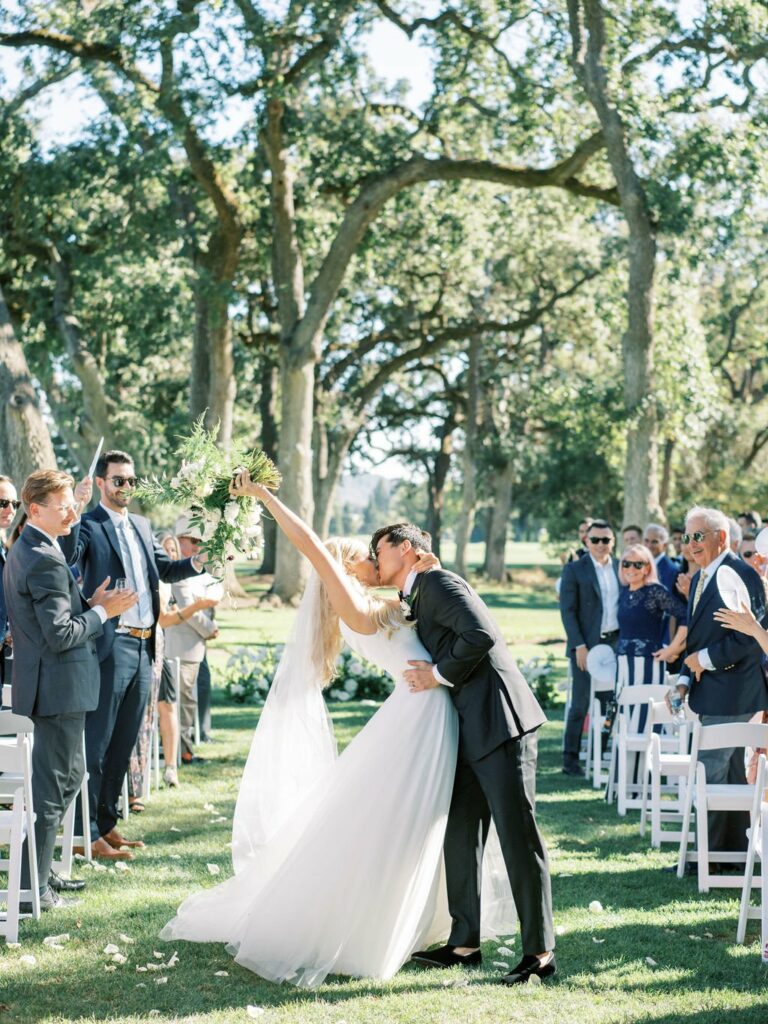 A bride and groom kiss at the end of a wedding aisle outdoors, surrounded by trees and guests who are clapping and smiling.