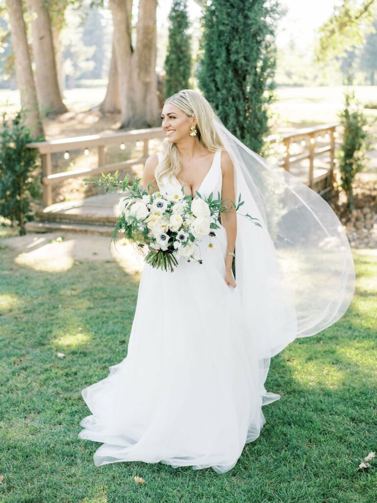 A bride in a white dress holds a bouquet of white and green flowers, standing in a garden with a small bridge and trees in the background. She has long, blonde hair and a flowing veil.