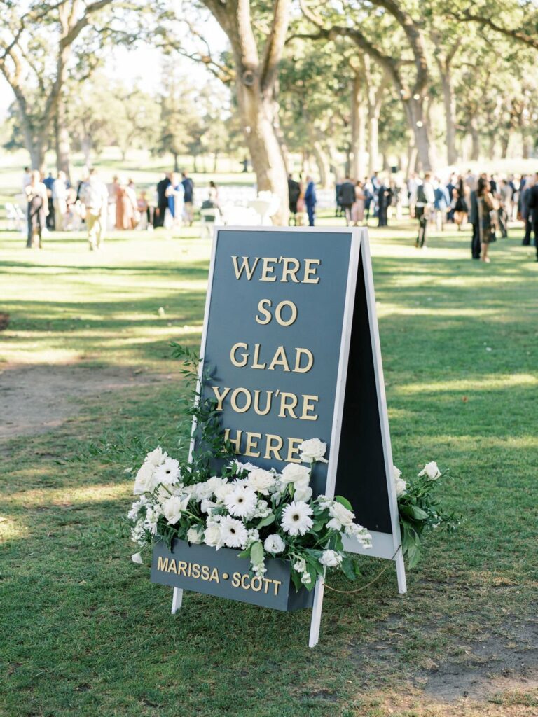 A double-sided sign with the text "We're so glad you're here" and "Marissa & Scott" stands on a grassy lawn adorned with white flowers, with people gathered in the background.