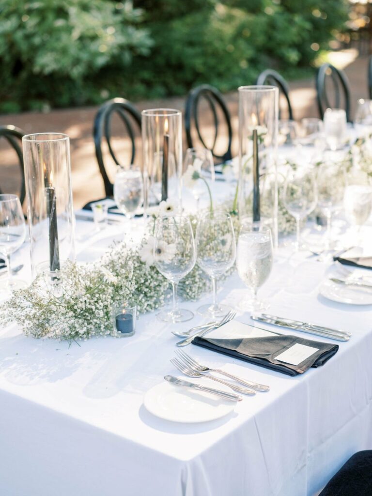 A long outdoor dining table is set with white linens, glassware, cutlery, baby's breath floral arrangements, and tall candles in hurricane vases. Black chairs are placed around the table.