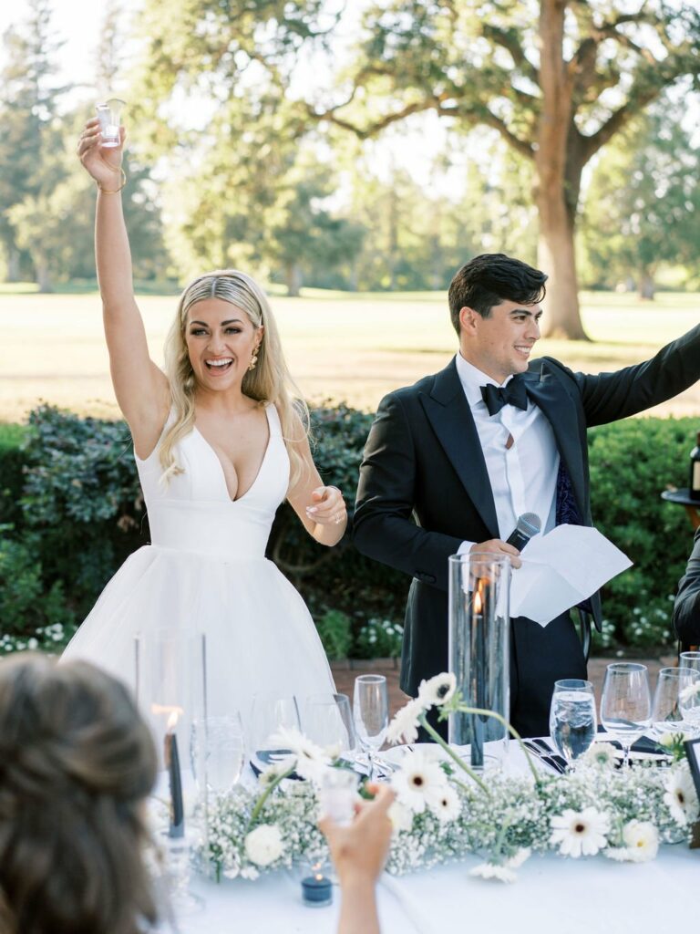 A bride in a white dress and a groom in a tuxedo stand outdoors, each raising a glass. They are smiling and appear to be making a toast at a wedding reception.