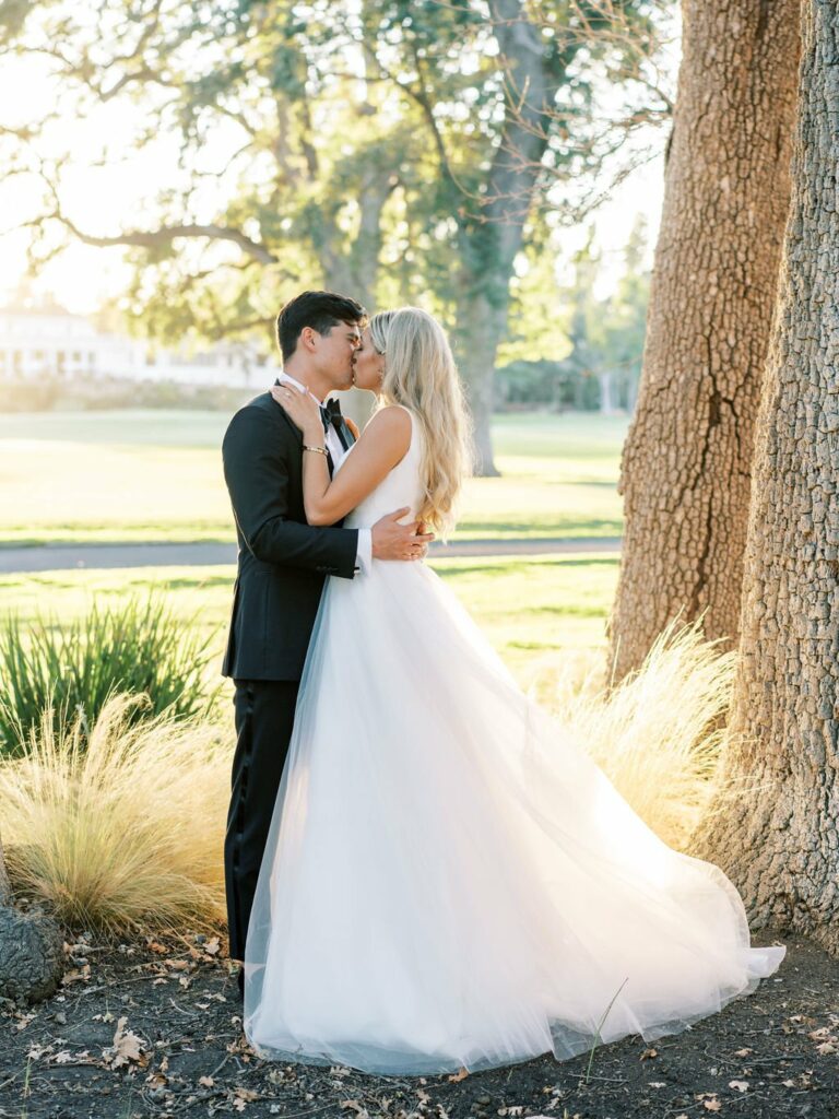 A bride in a white gown and a groom in a black suit kiss while standing close to a tree in a sunlit outdoor setting.