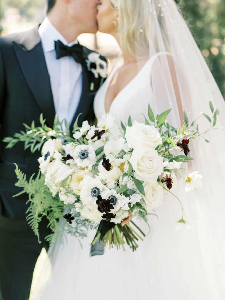 A bride and groom are kissing while holding a large bouquet of white and dark flowers. The bride is wearing a veil and a white dress; the groom is in a black tuxedo.
