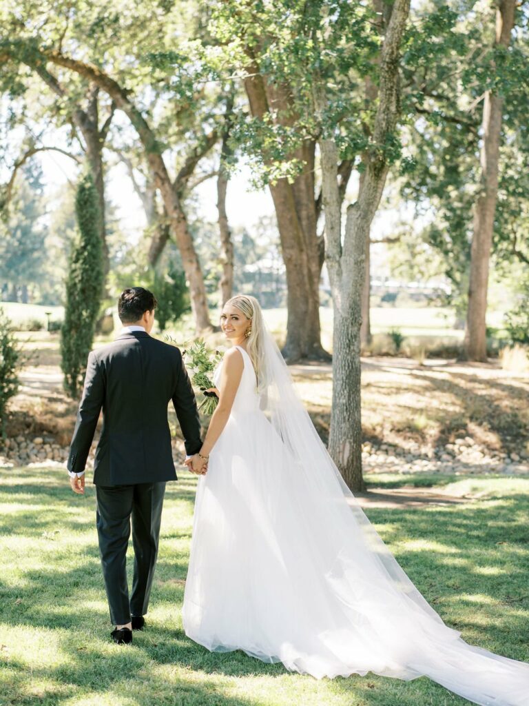 A couple dressed in wedding attire holds hands while walking outdoors on a grassy area surrounded by trees. The bride is wearing a long, flowing white dress, and the groom is in a dark suit.