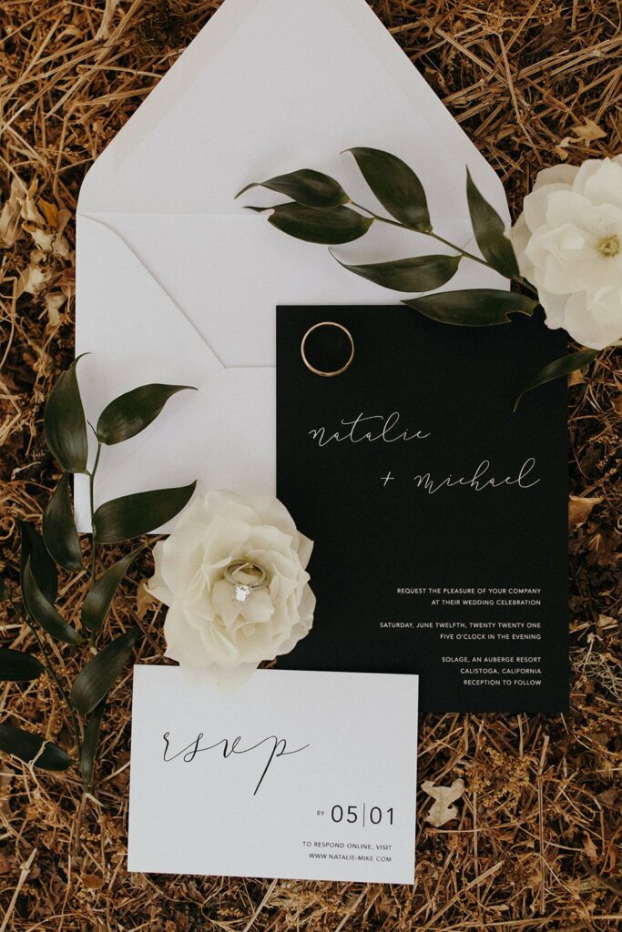 A wedding invitation set including an open white envelope, a black invitation card with "natalie + michael," an RSVP card, two white flowers, and greenery on a natural background.