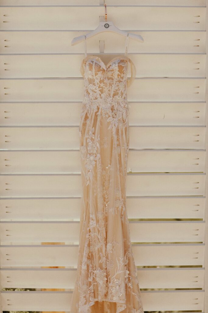A beige, strapless wedding dress adorned with lace detailing and floral embroidery is hanging on a white wooden slat wall.