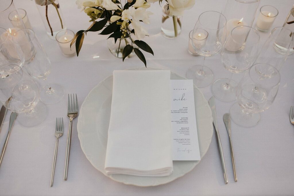 A white table setting featuring a folded napkin, a menu card, glassware, cutlery, and floral centerpiece, all arranged on a white tablecloth with lit candles.
