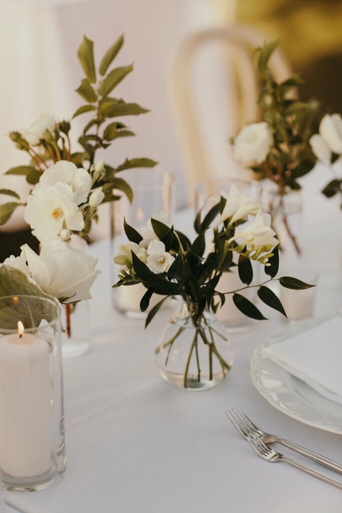 A dining table set with white flowers in glass vases, white candles, and white tableware, including a fork and knife on a white napkin, all arranged on a white tablecloth.
