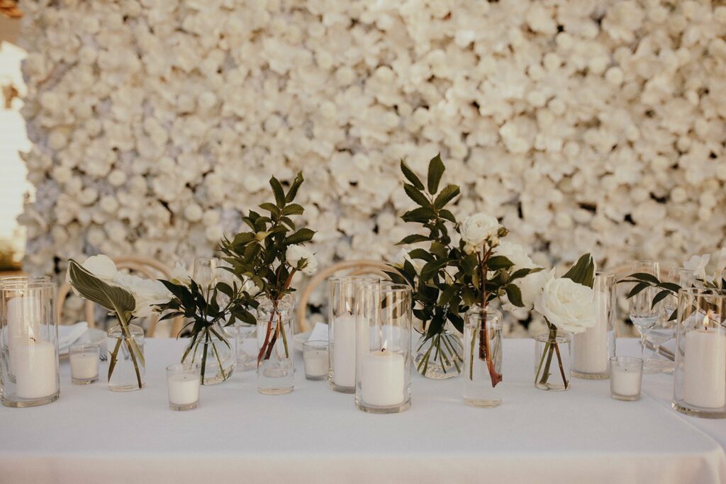 Table adorned with small glass vases holding white flowers and green leaves, alongside glass candle holders with white candles. White floral backdrop in the background.