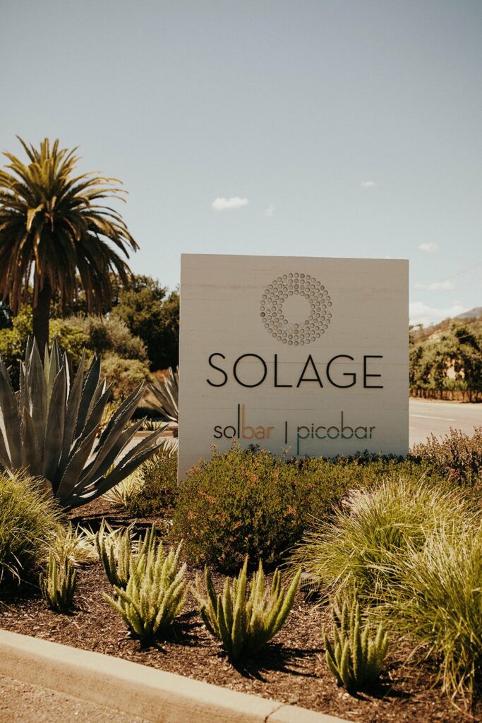 Sign for Solage resort featuring Solbar and Picobar, surrounded by lush landscaping and agave plants, with a palm tree in the background.