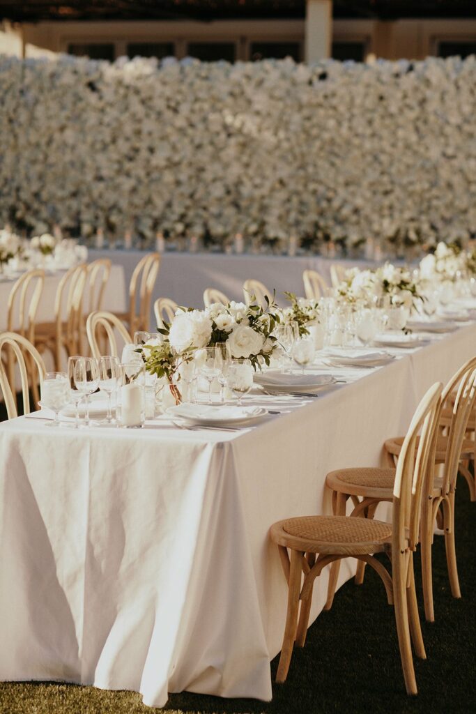 A long table with a white tablecloth is set with glassware, plates, and floral centerpieces. Wooden chairs are arranged on both sides, and a large wall of white flowers serves as a backdrop.