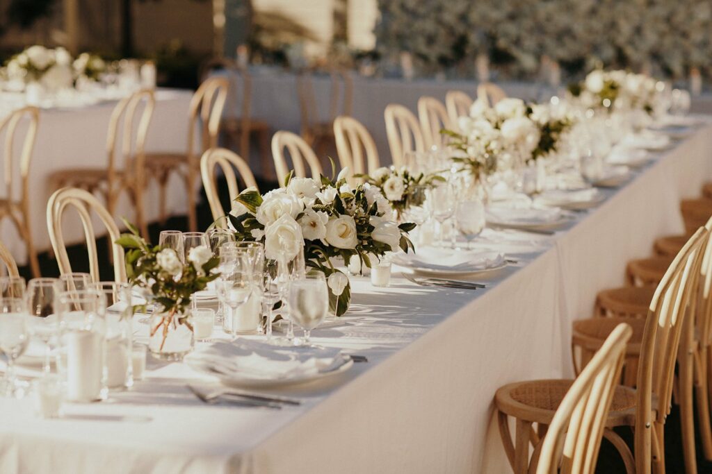 Long, elegantly set tables with white tablecloths and flower centerpieces, arranged for an outdoor event. Wooden chairs line both sides of the tables.