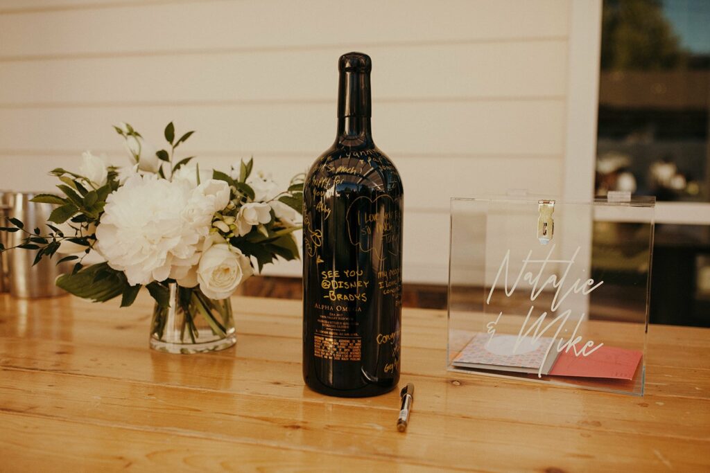 A large black wine bottle with gold writing sits next to a glass card box labeled "Natalie & Mike" and a bouquet of white flowers on a wooden table.