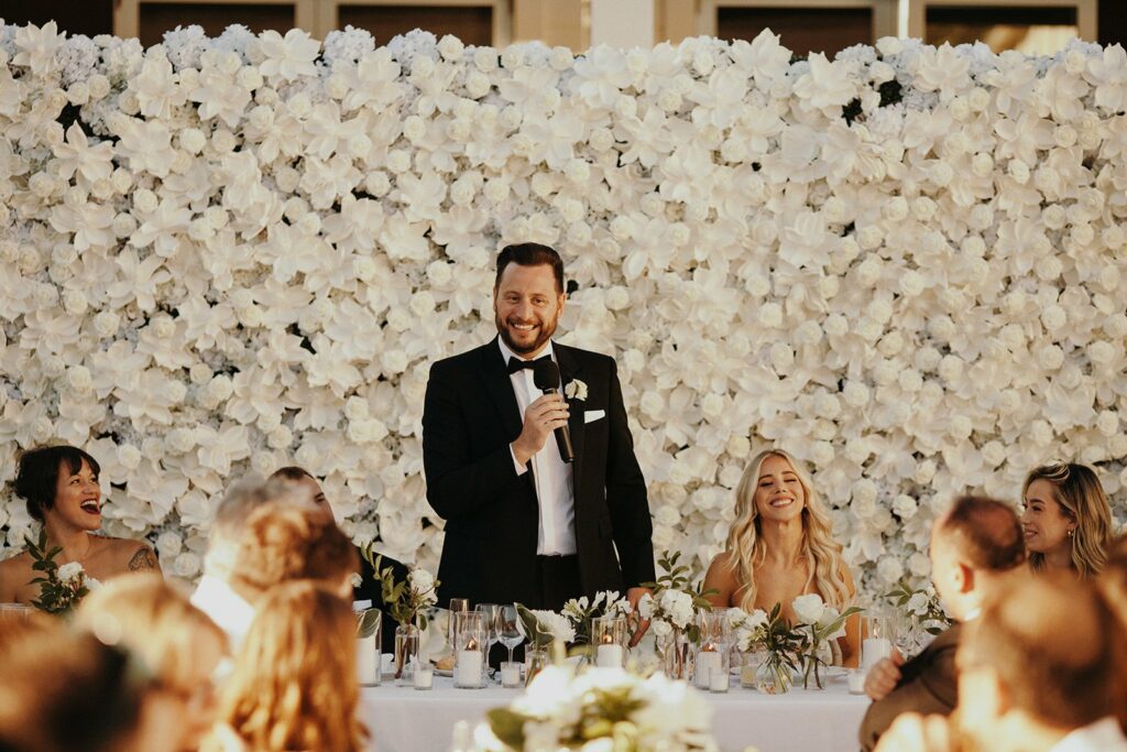 A man in a tuxedo is speaking into a microphone at a formal event, standing in front of a flower wall, with seated guests around him.